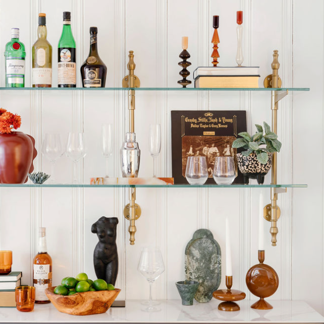 A beautiful custom shelving unit from Soil & Oak with glass shelves and brass hardware holds glasses, fruit, candles, and barware.