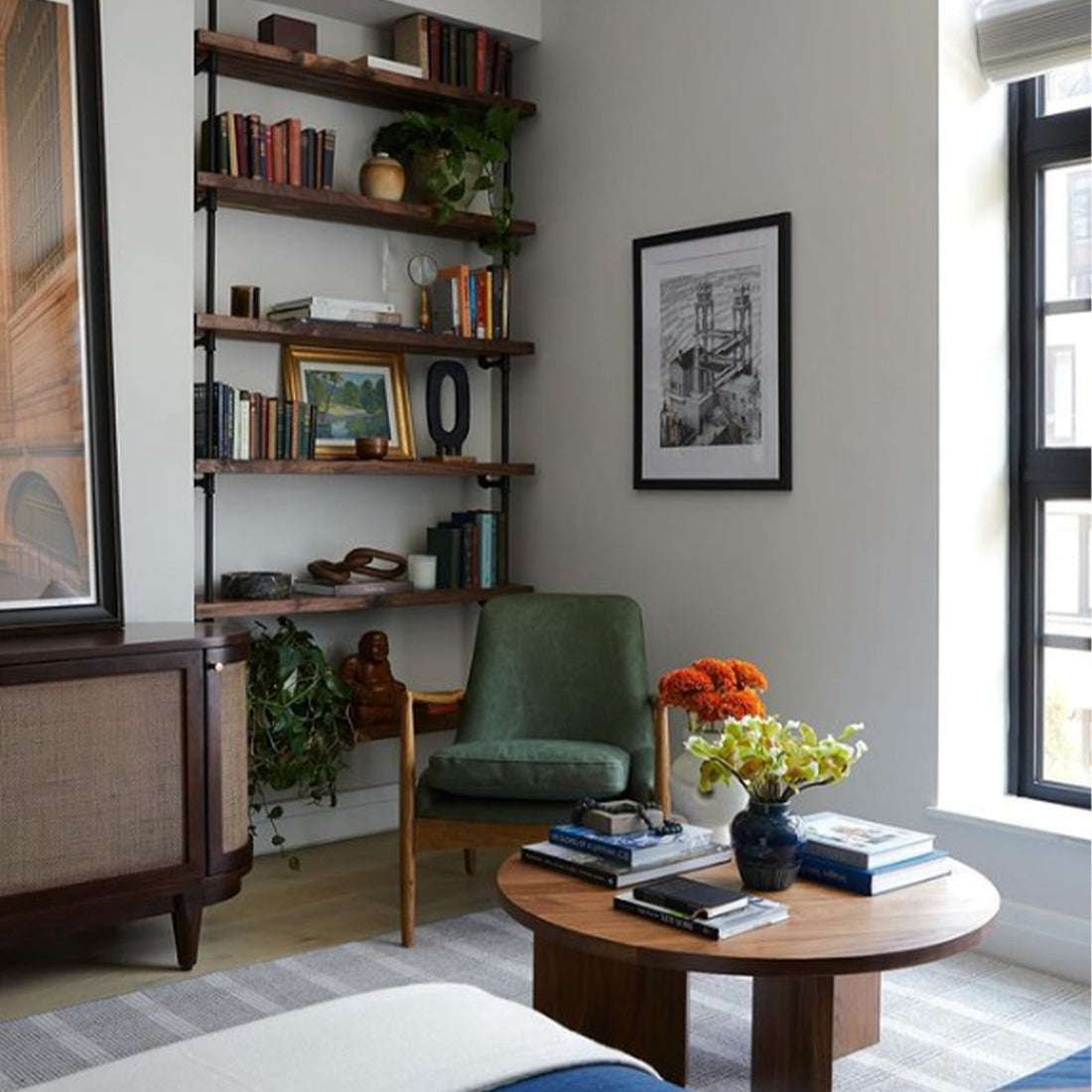 A custom Soil & Oak bookshelf with wood shelves and black piping is holding books, pictures, and plants in a modern NYC apartment. A simple green chair and circular wood table sit in front of it.
