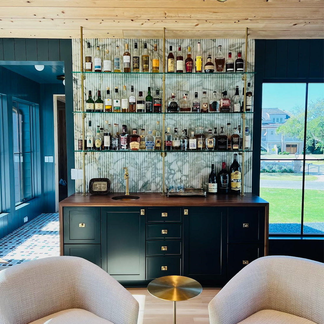 A stylish home bar featuring deep green cabinets, gold hardware, and glass shelves against a bold wallpaper.