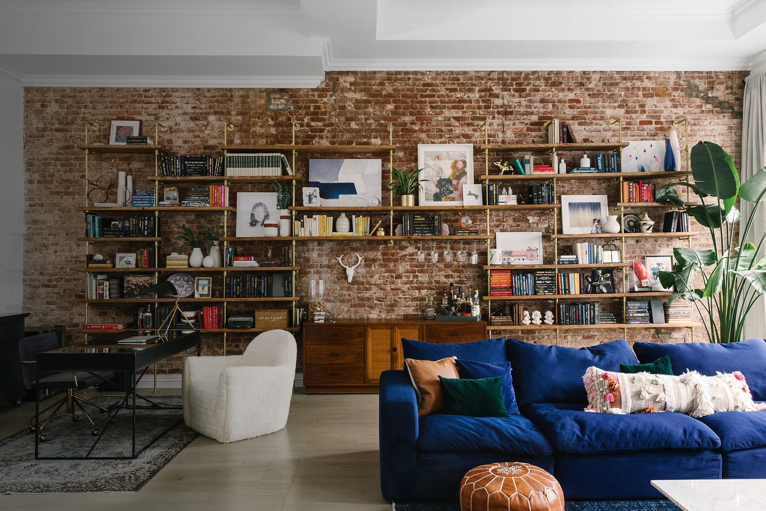 custom bookshelves with brass plated pipes and walnut shelves with a bar unit on a brick wall in a room designed by Tali Roth | Soil & Oak