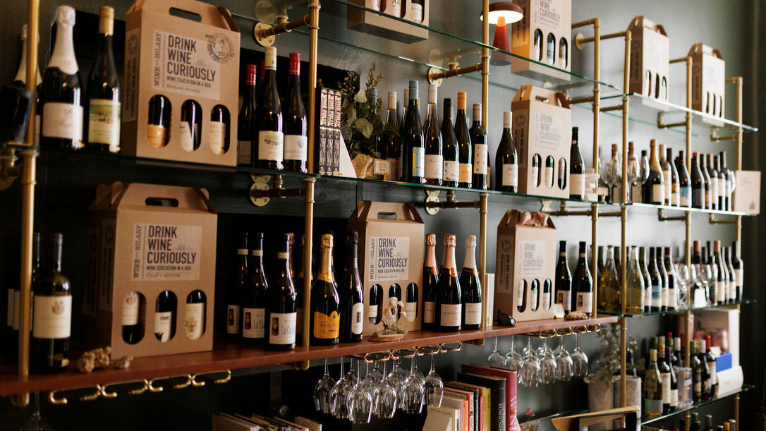 A beautiful wine shop has custom Soil & Oak shelves built with brass hardware and glass and wood shelves. They are willed with wine bottles, hanging glasses, and gift box sets.
