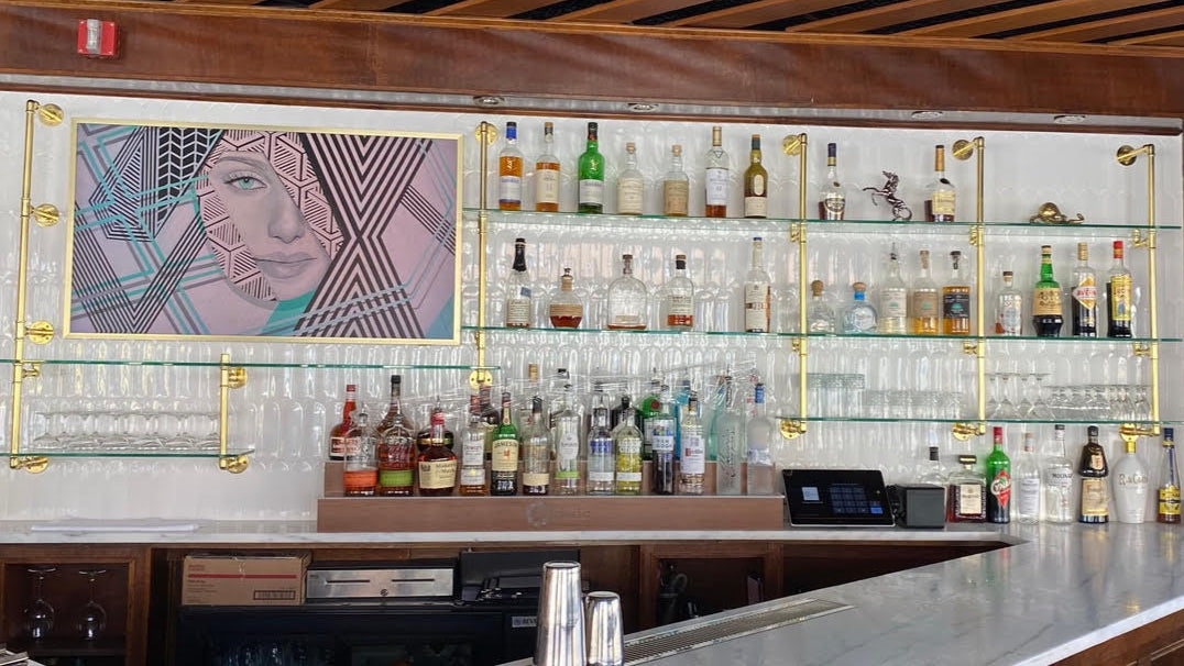 Custom bar built by Soil & Oak with shelves stocked with bottles, a painting of a woman's face on the wall, and a cash register.