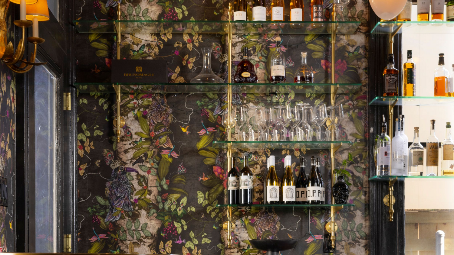 Bar interior with floral wallpaper, shelves with bottles, and chairs. Shelves built by Soil & Oak.