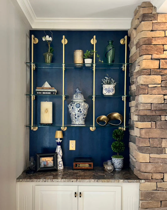 A custom shelving unit with brushed brass rails and glass shelves is installed on a dark blue wall above a marble countertop