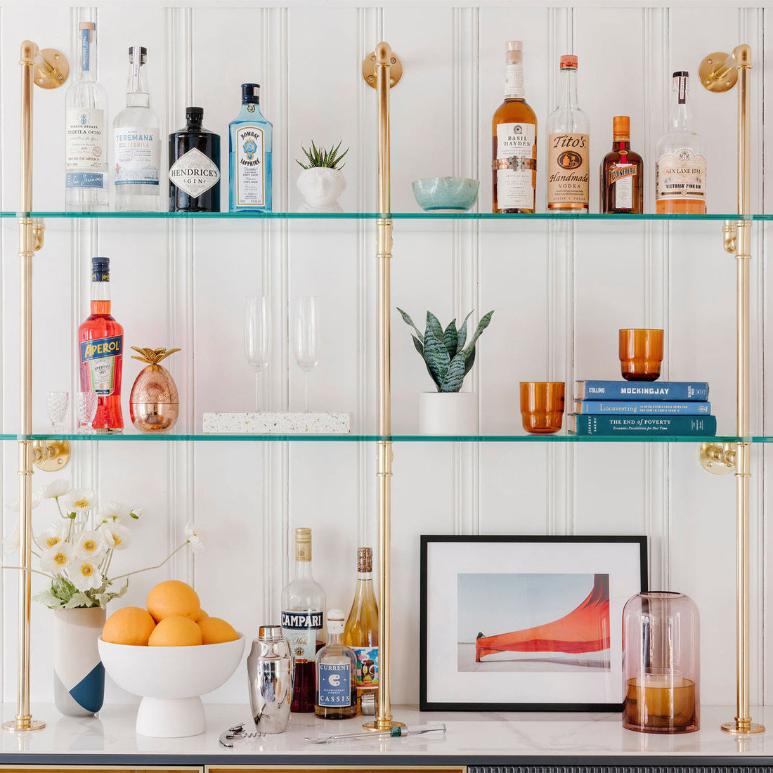 A stylish home bar featuring gold hardware and glass shelves is decorated with books and barware.