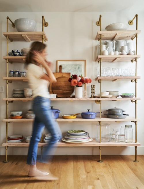 woman walking past a wall mounted shelves unit with brass pipes and maple butcher block shelves